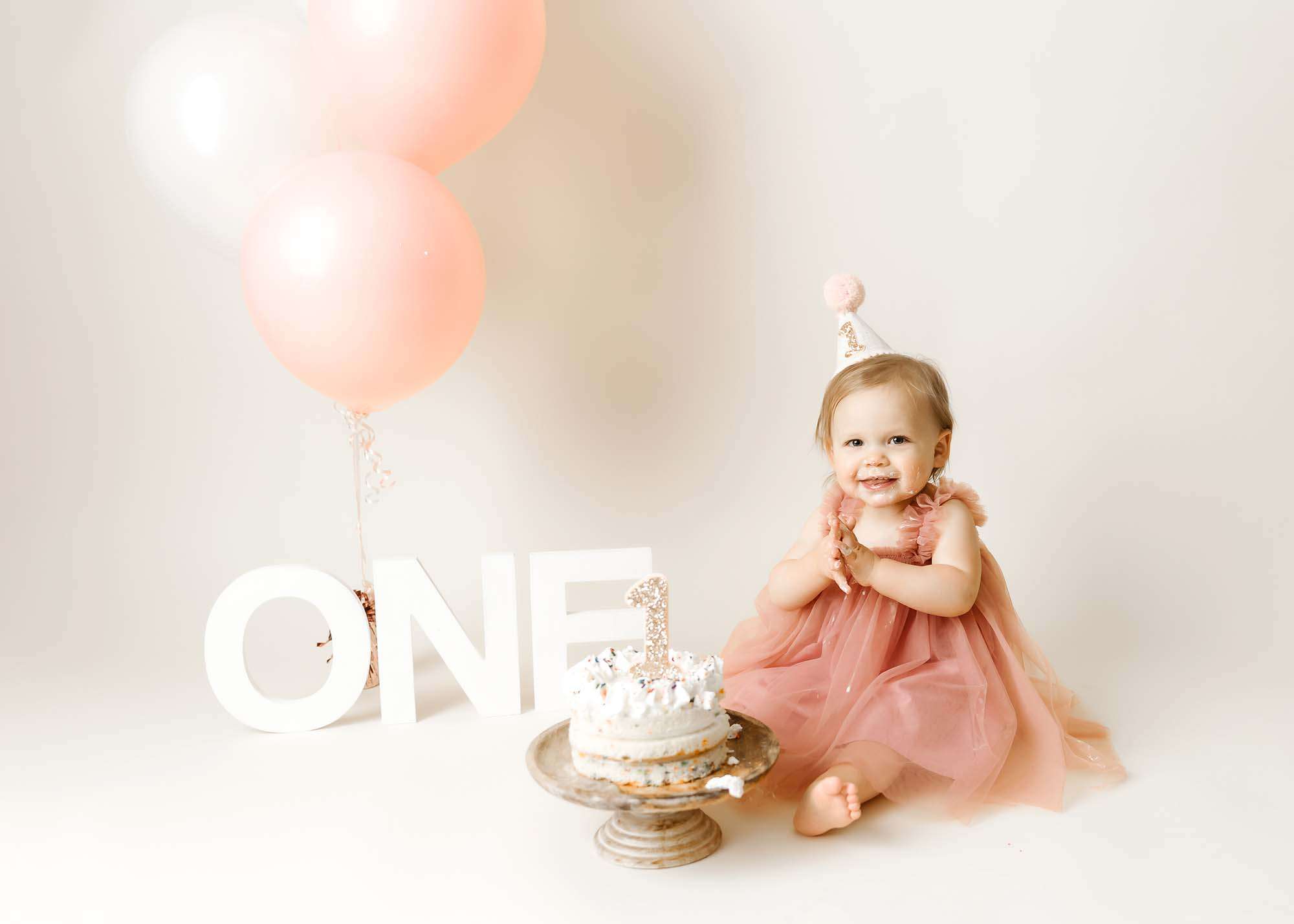 baby girl in pink dress with white frosted cake and pink balloons with minneapolis baby photographer