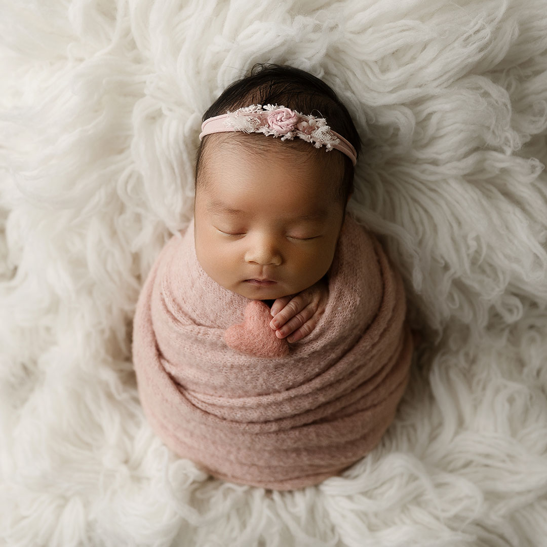 newborn girl wrapped in pink holding a pink heart on cream rug