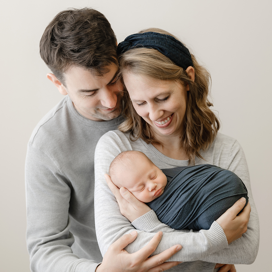 mom and dad in grey holding newborn boy wrapped in navy blue in minneapolis mn
