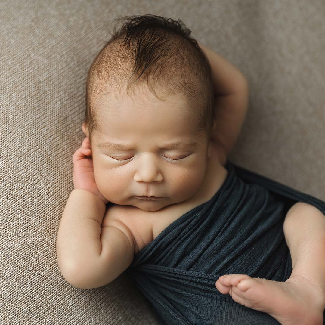 newborn boy wrapped in blue laying with hands behind head in newborn studio minneapolis