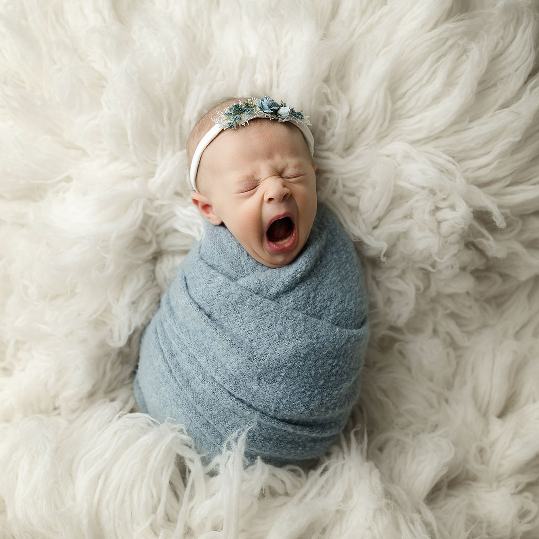 newborn girl wrapped in blue lying on cream rug yawning in minneapolis newborn studio