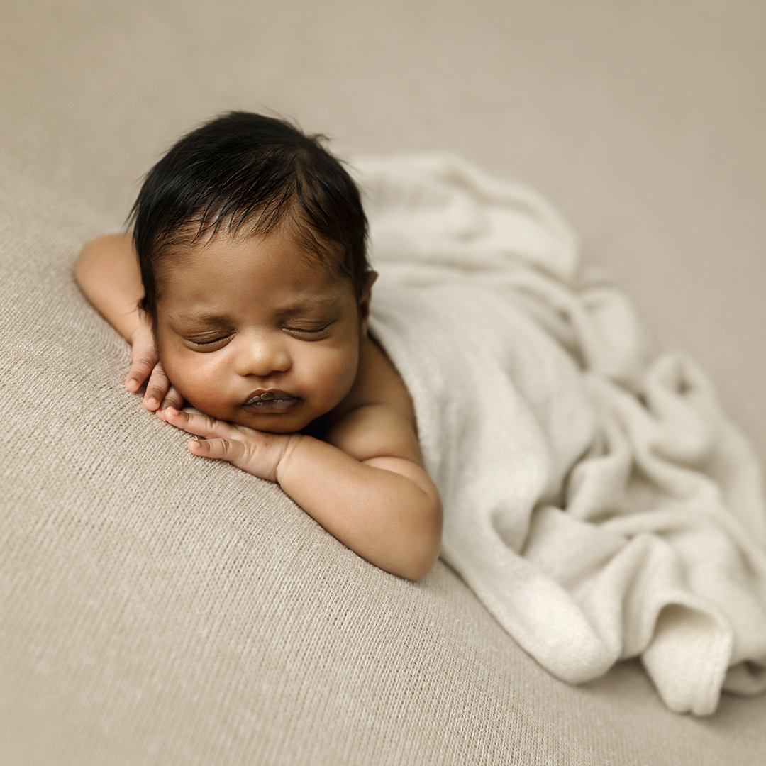 Newborn lying on brown blanket covered in cream wrap with chin resting on hands