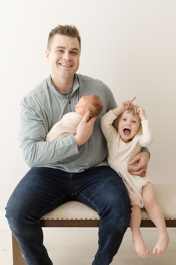 dad holding newborn sitting with toddler on bench in newborn photography studio