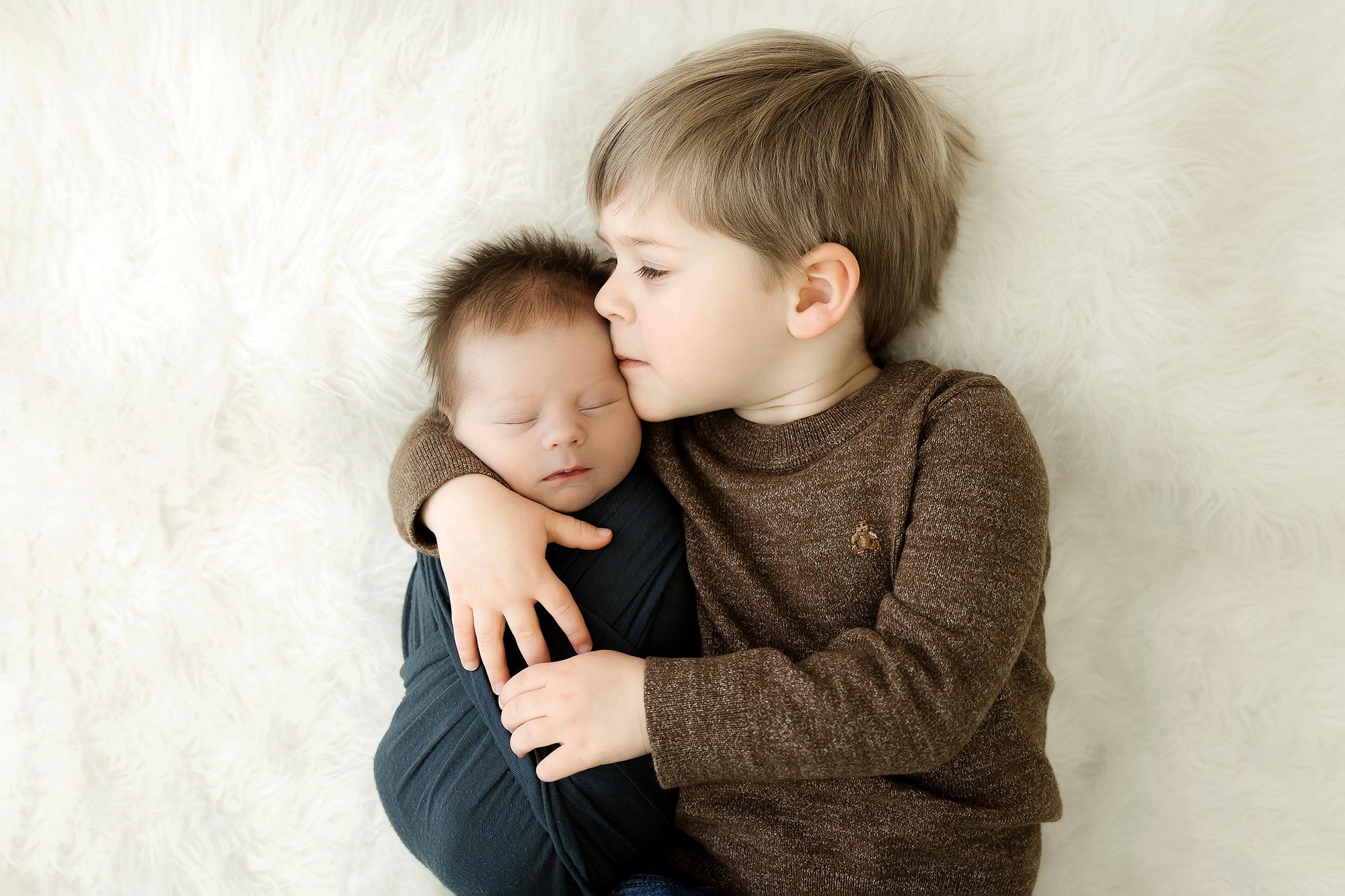 older brother kissing newborn brother lying on white rug in minneapolis portrait studio