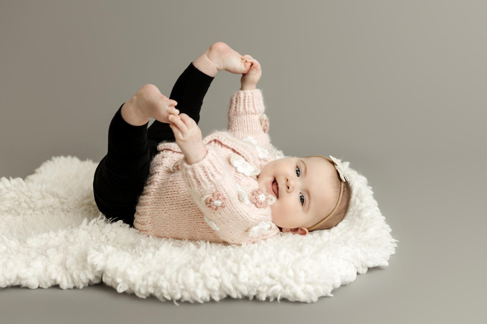 6 month old baby girl lying on back grabbing her toes in baby portrait studio