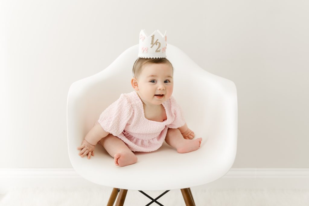 6 month baby girl with crown sitting on white chair in plymouth baby photography studio